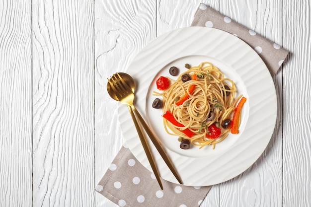 Espaguetis alla caponata de pasta integral con tomates, aceitunas, pimiento rojo dulce y alcaparras en un plato blanco sobre una mesa de madera blanca con textura, endecha plana