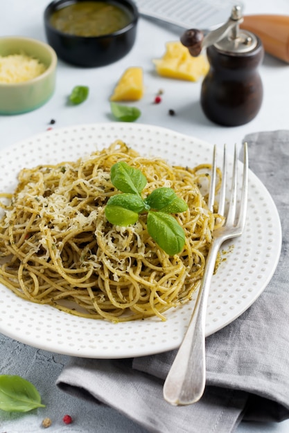 Espaguetis de pasta con salsa pesto, albahaca y queso parmesano sobre una placa de cerámica blanca y fondo gris de hormigón o piedra. Plato tradicional italiano.