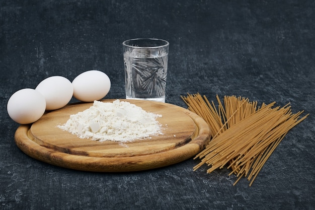Espaguetis secos, harina sobre una tabla de madera para cortar, huevos de gallina y un vaso de agua
