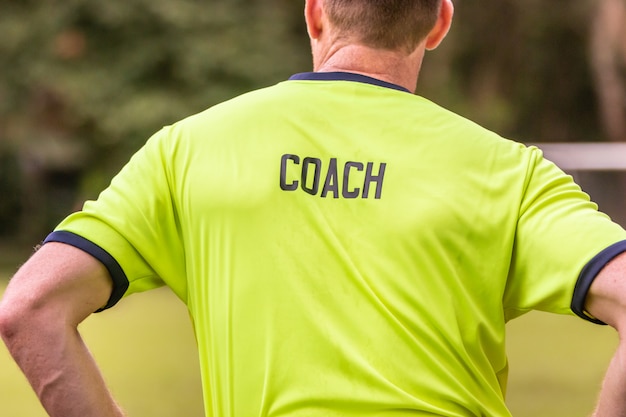 La espalda del entrenador masculino, con la palabra COACH escrita en su camisa, en un campo al aire libre