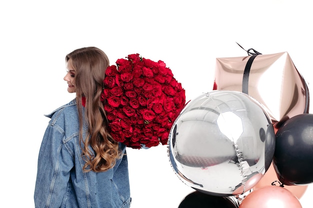 Espalda de mujer con chaqueta de jeans azul sosteniendo un gran ramo de rosas en su hombro Chica morena con cabello largo recibiendo regalos en cumpleaños Elegante dama de pie cerca de globos dorados, plateados y negros