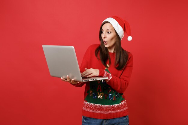 Espantada com a jovem garota de chapéu de Natal, parecendo surpresa, trabalhando digitando no computador laptop pc isolado sobre fundo vermelho. Conceito de festa de feriado de celebração de ano novo feliz 2019. Simule o espaço da cópia.