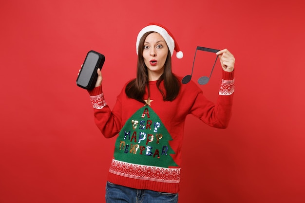 Espantado com a jovem garota de Papai Noel segurando a nota musical, alto-falante portátil sem fio bluetooth música isolado em fundo vermelho brilhante. Conceito de festa de feriado de celebração de ano novo feliz 2019. Simule o espaço da cópia.