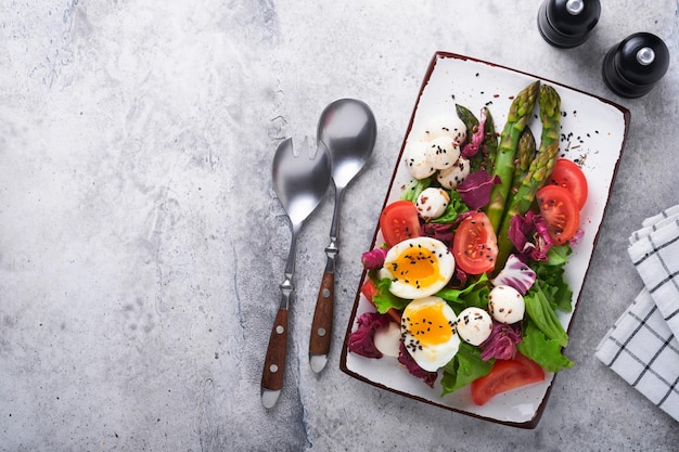 Espargos tomate alface mussarela gergelim preto óleo de linhaça salada de azeitona e ovo cozido macio na placa cerâmica retangular no fundo da mesa de concreto cinza Conceito de comida de dieta saudável Vista superior