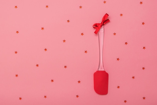 Espátula de cozinha vermelha brilhante decorada com fita vermelha em fundo rosa com estrelas. Utensílios de cozinha em fundo festivo. Postura plana.