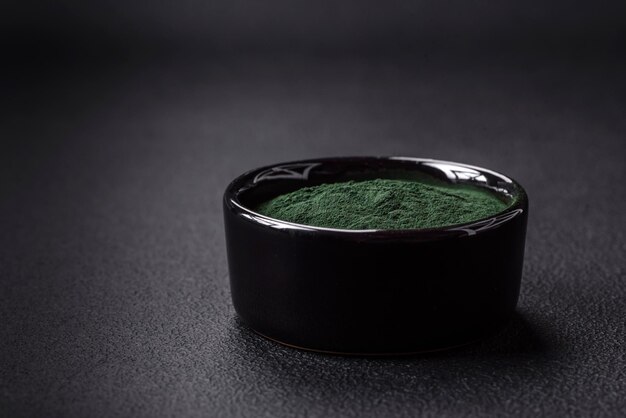 Foto especia de espirulina de color verde en forma de polvo en un recipiente negro sobre un fondo de hormigón oscuro