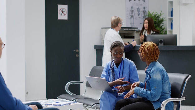 Especialista en consultar a un paciente afroamericano en el vestíbulo de recepción de la sala de espera, usando una computadora portátil para explicar el tratamiento de la enfermedad y la diangosis. Haciendo consulta de salud y examen con mujer.