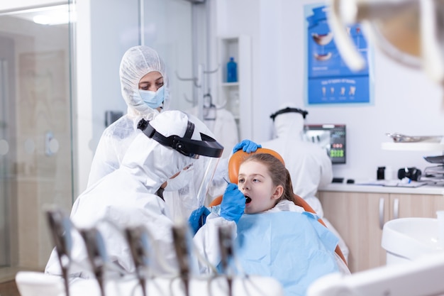 Especialista dental en traje de ppe que comprueba la cavidad del niño durante la visita al dentista. Dentista en traje de coronavirus con espejo curvo durante el examen de los dientes del niño.