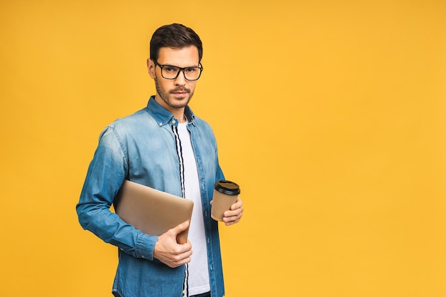 Especialista em negócios confiante, jovem e bonito homem barbudo em casual segurando laptop e sorrindo em pé sobre fundo amarelo isolado