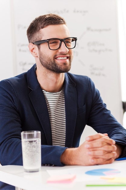 Especialista em negócios confiável. Homem jovem confiante em roupas casuais inteligentes, olhando para o lado e sorrindo enquanto está sentado em seu local de trabalho com um quadro branco ao fundo