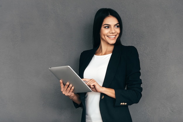 Especialista em negócios confiável. Mulher jovem e atraente em moda casual inteligente, trabalhando em um tablet digital e sorrindo em pé contra um fundo cinza