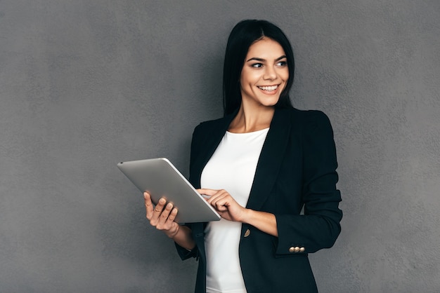 Especialista em negócios confiável. mulher jovem e atraente em moda casual inteligente, trabalhando em um tablet digital e sorrindo em pé contra um fundo cinza