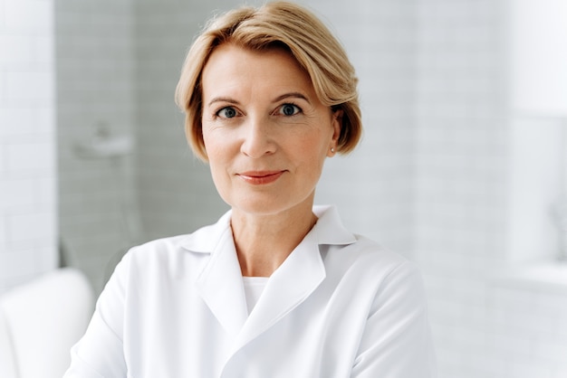 Especialista hábil. Retrato de cosmetóloga madura en bata de laboratorio blanca mirando a la cámara con una sonrisa mientras posa en su clínica