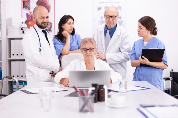 Especialista médico maduro que informa a su equipo médico en la sala de conferencias usando la computadora portátil. Terapeuta experto de la clínica hablando con colegas sobre la enfermedad, profesional de la medicina