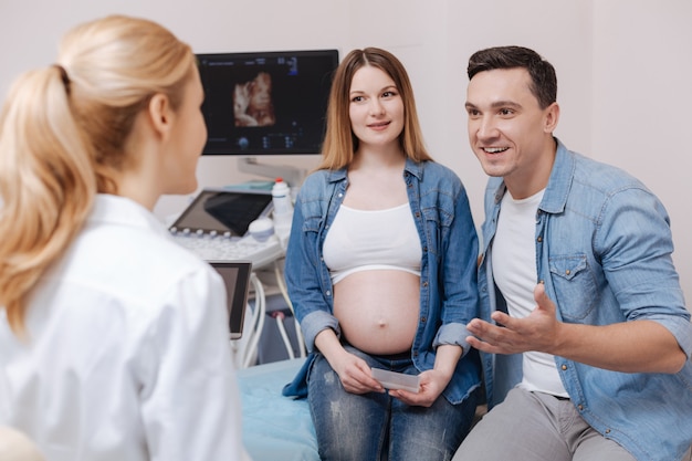 Especialista médico profesional involucrado encantado que trabaja en la clínica y conversa con una pareja joven mientras se habla de un parto futuro