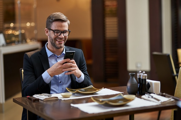 Especialista en negocios masculino sonriendo mientras sostiene el teléfono con ambas manos frente a su rostro en la mesa del restaurante