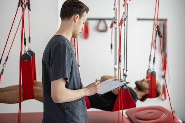 Especialista en rehabilitación que examina a un paciente masculino antes del tratamiento activo con correas de suspensión