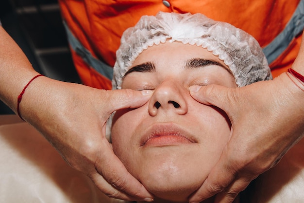 El especialista en spa está haciendo un masaje facial a una joven con un gorro médico tratamientos de belleza cerrados