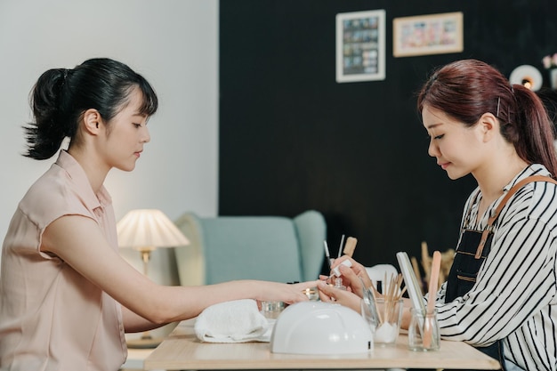 especialista en uñas haciendo manicura para el cuidado de las uñas para el cliente en la mesa en el estudio. la joven oficinista después del trabajo disfruta del tiempo libre para pintar uñas de moda en el salón de belleza. manicurista en delantal sonriendo en taller