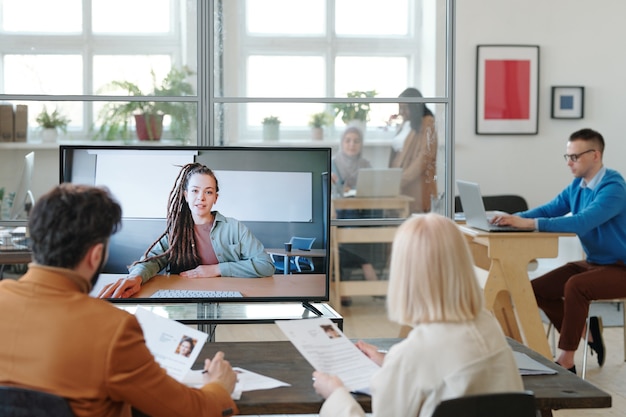 Especialistas de RH sentados com currículos à mesa e ouvindo a apresentação do candidato a emprego por meio do aplicativo de bate-papo por vídeo no computador de um escritório moderno