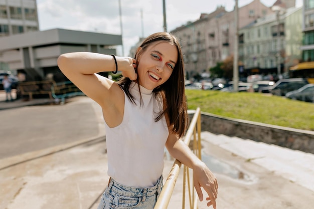 Espectacular mujer encantadora con el pelo oscuro vestida con una camiseta blanca posando en la cámara en el centro de la ciudad a la luz del sol