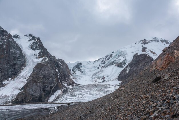 Espectacular paisaje con caída de hielo glaciar en una gran cadena montañosa nevada con un pináculo rocoso afilado bajo un cielo gris nublado Gran cierre de glaciar en gran altitud Paisaje sombrío en montañas nubladas