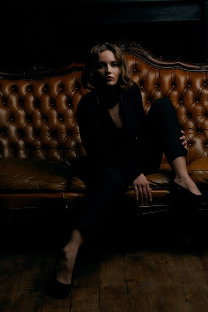 Espectacular retrato de estudio de sensual chica morena con maquillaje perfecto viste traje negro sentado en un sofá marrón vintage