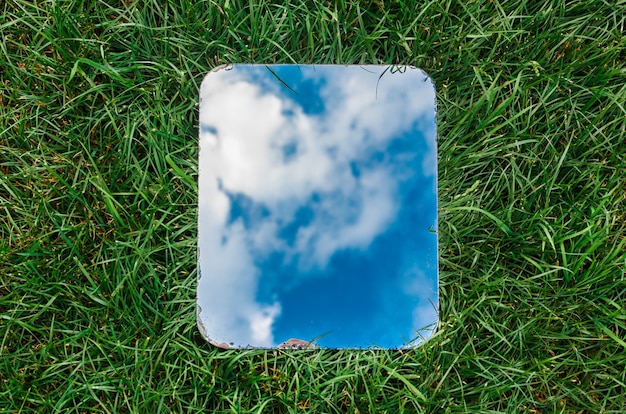 Espejo rectangular que refleja el cielo azul con nubes sobre la hierba verde.