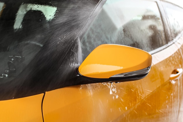 Espelho lateral do carro amarelo sendo lavado em autoatendimento, pulverização de água com alta pressão, gotas voando ao redor.