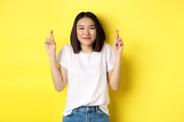 Esperançosa garota asiática sorrindo otimista, sentindo-se com sorte, cruzando os dedos e fazendo pedido, olhando para cima sonhadora, em pé sobre um fundo amarelo