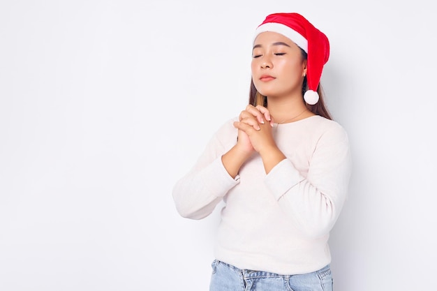 Esperançosa jovem asiática com chapéu de Natal mantém as palmas das mãos pressionadas em gesto de oração isolado no fundo branco