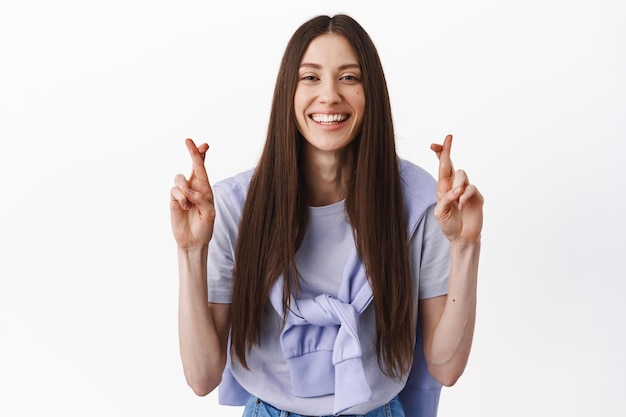 Esperançosa mulher caucasiana cruza os dedos para dar sorte, sorrindo e olhando para a câmera, fazendo um desejo, esperança pela realização de um sonho, antecipando boas notícias, em pé sobre um fundo branco