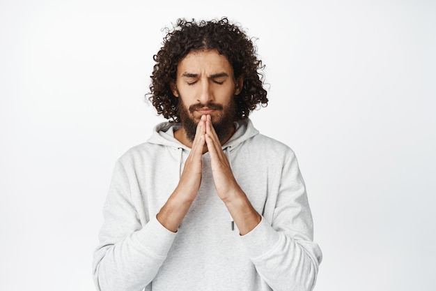 Esperançoso homem hispânico preocupado rezando suplicando a Deus esperando por algo em pé sobre fundo branco