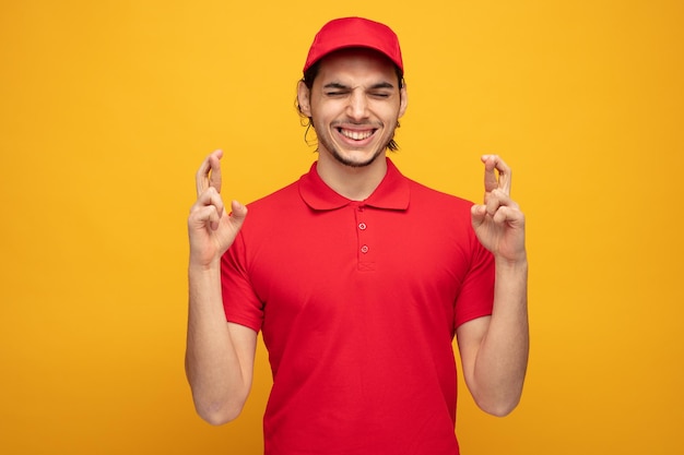 Esperançoso jovem entregador vestindo uniforme e boné fazendo desejo com os olhos fechados, mantendo os dedos cruzados isolados no fundo amarelo