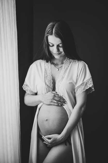 Esperando bebé Embarazada mujer sexy de pie cerca de la ventana en casa y las manos abraza el vientre redondo estómago cerrar nueve meses Baby Shower Concepto de maternidad Lugar para texto Foto en blanco y negro