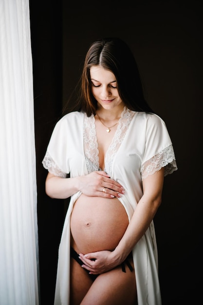 Esperando bebé Embarazada mujer sexy de pie cerca de la ventana en casa y las manos abraza un vientre redondo estómago cerrar nueve meses Baby Shower Concepto de maternidad