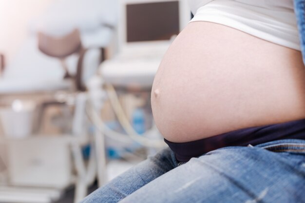 Esperando cita médica. Joven mujer embarazada pacífica sentada en el gabinete de ginecología y esperando al médico mientras se prepara para la cita
