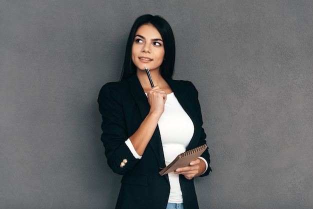 Esperando inspiración. Mujer joven atractiva en ropa de sport elegante que sostiene la pluma y el cuaderno y que mira pensativa mientras está de pie contra el fondo gris