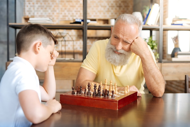 Esperando pacientemente. Agradável homem sênior jogando xadrez com seu adorável neto pré-adolescente e olhando para ele com um sorriso, esperando seu próximo movimento