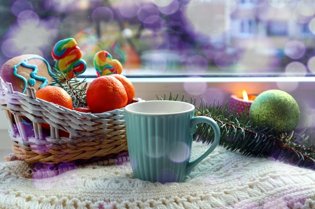 Esperando pelo feriado: uma caneca de chá em um suéter branco de tricô, tangerinas, uma vela acesa, um galho de abeto no parapeito da janela, bokeh