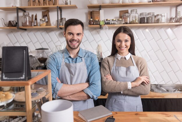 Esperando por clientes. casal jovem encantador sorrindo e cruzando as mãos no peito em pé atrás do bar.