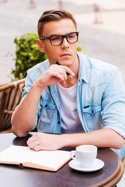 Esperando por inspiração. Jovem pensativo tocando seu queixo com uma caneta e desviando o olhar enquanto está sentado em um café na calçada com um bloco de notas sobre a mesa