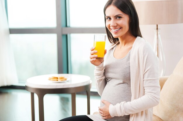 Esperando por uma nova vida. Mulher jovem e bonita grávida segurando um copo com suco de laranja e sorrindo