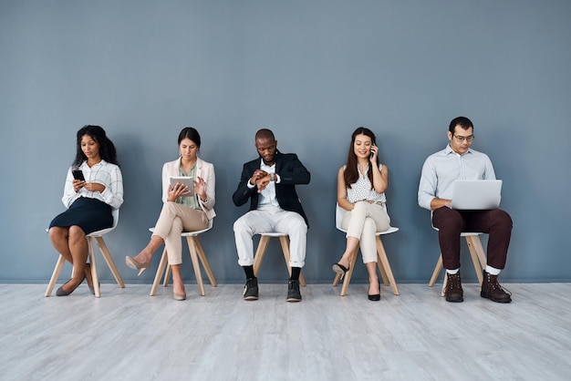 Esperando sua vez por uma chance de progressão na carreira Foto de um grupo de empresários usando dispositivos digitais enquanto estão sentados na fila contra um fundo cinza