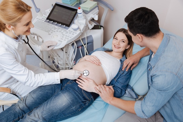 Esperando tener un niño sano. Carismático joven esposo alegre sentado en la clínica y apoyando a la esposa mientras el médico prueba el examen del vientre embarazado