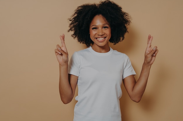 Esperanza femenina afroamericana aislada sobre fondo beige studio cruzando los dedos haciendo deseo