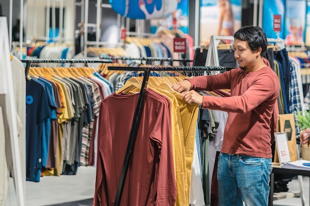 Esperto homem asiático com barba escolher roupas em loja de roupas no centro comercial