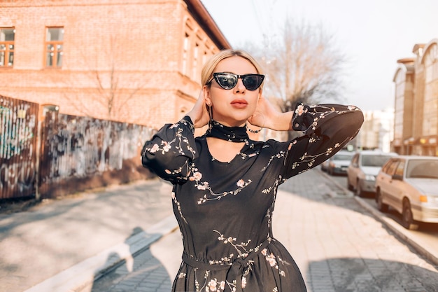 Espetacular modelo feminino caucasiano loiro em vestido preto estampado floral e óculos de sol tocando o rosto olhando para a câmera com sorriso em pé perto da velha porta de madeira. estilo de vida, conceito de viagem.