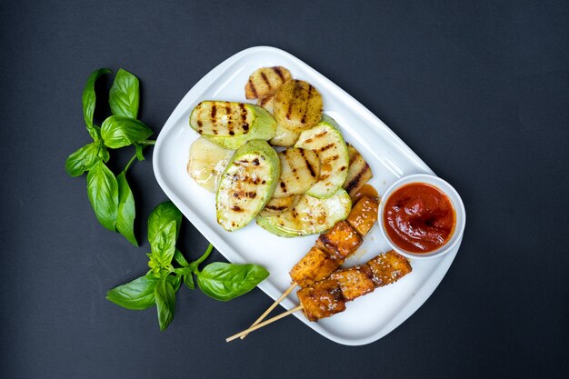 Espetada de tofu grelhado em um prato branco. Fundo preto. Folhas frescas de manjericão.