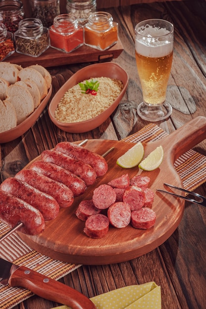 Espeto de salsicha grelhada em tábua de madeira com cerveja, pão e farofa churrasco brasileiro